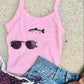 Pink tank top with Rainbow trout, sunglasses graphic, blue jeans, and brown sandals on a beach surface