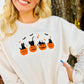 A smiling blonde woman wearing a white long-sleeve sweatshirt with a Halloween graphic of three black cats in orange pumpkins, posed outdoors in front of a tree trunk with warm string lights.