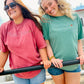 Two women smiling outdoors while wearing graphic t-shirts. The woman on the left wears a dusty rose pink t-shirt with 'Roll Tide' in white script, and the woman on the right wears a sage green t-shirt with 'Heritage Warriors' in white script.