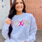 Woman wearing a white sweatshirt with heart and X puff glitter, standing in front of a brick wall.