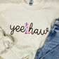 A tan embroidered sweatshirt with the text 'YEE HAW' on it, displayed on a surface with blue jeans and other clothing items.