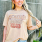 Woman wearing a beige t-shirt with text and denim shorts standing against a concrete wall Praying moms club.