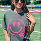 A female model wearing a gray t-shirt with a neon pink smiley face graphic on it, paired with denim shorts and mascot name, standing on an athletic field.