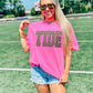 A person wearing sunglasses and a neon pink t-shirt with the word 'TIDE' embroidered in brown on the front, standing on an athletic field.