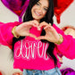 Woman in a pink sweatshirt with 'LOVER' text, making a heart shape with her hands, surrounded by heart-shaped balloons.