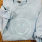 A light blue sweatshirt with an embroidered lettering of a smiling face on the chest, displayed on a woven mattress.