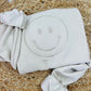 A beige embroidered sweatshirt with a happy face design on the front, displayed on a woven mat.
