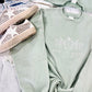 A green embroidered sweatshirt with 'wildflowers' text and a floral pattern, displayed flat with a pair of white sneakers and a denim jacket in the background.