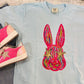 A light blue colored t-shirt with a graphic print of an eye-catching, colorful Easter bunny on it, displayed flat with a pair of pink sneakers and a white shoe on top of it.