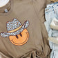 A brown short-sleeved graphic tee with a leopard wearing a cowboy hat and the text 'HAPPY' on it, displayed on a surface with jeans and shoes.