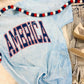 A blue colored graphic tee with the text 'AMERICA' printed in red and white, displayed on a faux fur surface with jeans and a decorative lei in the background.