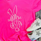 Pink t-shirt with 'Happy Easter' bunny design on a fluffy surface with sneakers.