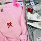 The image displays a light pink embroidered sweatshirt with the phrase "BE MINE" and a red ribbon design, presented next to a pair of blue denim jeans. The sweatshirt features decorative red heart-shaped tags and embroidered stars, enhancing its Valentine's Day-themed aesthetic.