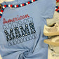 A blue t-shirt with the text 'American MAMA' printed on it, presented on a flat surface with decorative items around it.