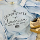 A light blue t-shirt with the text 'The United States of America Home of the Brave' printed on it, displayed flat with a pair of beige shoes at the bottom of the frame.