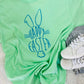 Light green t-shirt with 'Happy Easter' design on a fluffy white surface with sneakers.