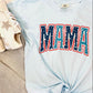 A blue curved-neck t-shirt with the word 'MAMA' and a star graphic in red and blue colors, displayed flat with a pair of white sneakers and a furry white blanket in the background.