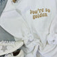 A white retro wave embroidered sweatshirt with the text 'you're so golden' on the front, displayed on a furry surface with a pair of sneakers and a star-shaped brooch.
