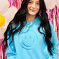 A person wearing a blue sweatshirt with a happy face embroidery on the front, posing against a pink glittery background with balloons.