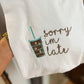 A white fabric with embroidered text 'Sorry I'm late' and a stylized iced coffee design held in a hand.