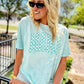 A woman wearing a light blue graphic tee with a checkered pattern, paired with shorts and sunglasses, standing near a metal railing.
