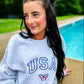 A person sitting by a poolside wearing an ash grey sweatshirt with the embroidered letters 'USA' and a butterfly logo on the front.