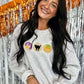 A woman smiling, wearing a long-sleeved gray sweatshirt with orange and black embroidered halloween cookies, and a sparkly background.