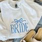 A white hoodie with the word 'Bride' embroidered on the front, paired with blue jeans and beige flats on a faux furry surface.