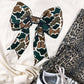 White sweatshirt with a large camouflage bow, leopard print pants, and Adidas sneakers on a white background.