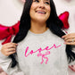 A woman smiling and posing with a gray sweatshirt that has the word 'lover' written in neon pink and a pink bow graphic.