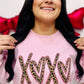 A person wearing a light pink sweatshirt with the word 'XOXO' embroidered in a leopard print style on the front, surrounded by heart-shaped decorations.