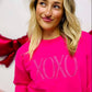 A woman wearing a hot pink embroidered sweatshirt with the text 'XOXO' on it, posing against a background of red and white balloons and decorations.