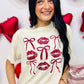 A person wearing a beige t-shirt with a graphic print of three stylized lips and bows in red and white colors, standing against a backdrop of Valentine's Day-themed decorations including balloons and hearts.