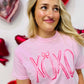 A woman wearing a pink t-shirt with the text 'XOXO' and a pink bow design on it, standing in a room with balloons and decorative hearts.