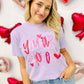 A person wearing a purple graphic tee with the text 'you're loved' and heart doodles, posing with a background of red and white heart-shaped decorations.
