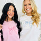 Two individuals standing next to each other, one wearing a pink and the other wearing a white embroidered bow sweatshirt.