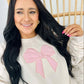 A woman smiling and posing in a beige sweatshirt with a pink bow applique on the front.