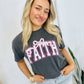 A woman wearing a gray t-shirt with the word 'Faith' and a pink bow on it, standing with one hand against a white background.