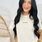 A woman wearing a white short-sleeved t-shirt with the word 'Mom' embroidered on the front, standing in a room with a wooden rocking chair and a watch on her wrist.