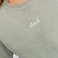A person wearing a sandstone-colored t-shirt with the word 'DAD' embroidered on the front.