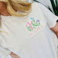 A woman wearing a white short-sleeved t-shirt with embroidered floral design and the word 'mama' on the front.