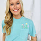 A woman wearing a chalky mint green t-shirt with a floral pocket design and embroidered lettering on the front, smiling against a white background.