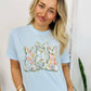 Woman wearing a light blue t-shirt with a colorful graphic design, standing indoors.