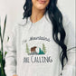 A woman smiling, wearing an ash grey embroidered sweatshirt with the text 'THE MOUNTAINS ARE CALLING' and an image of a bear in a forest.
