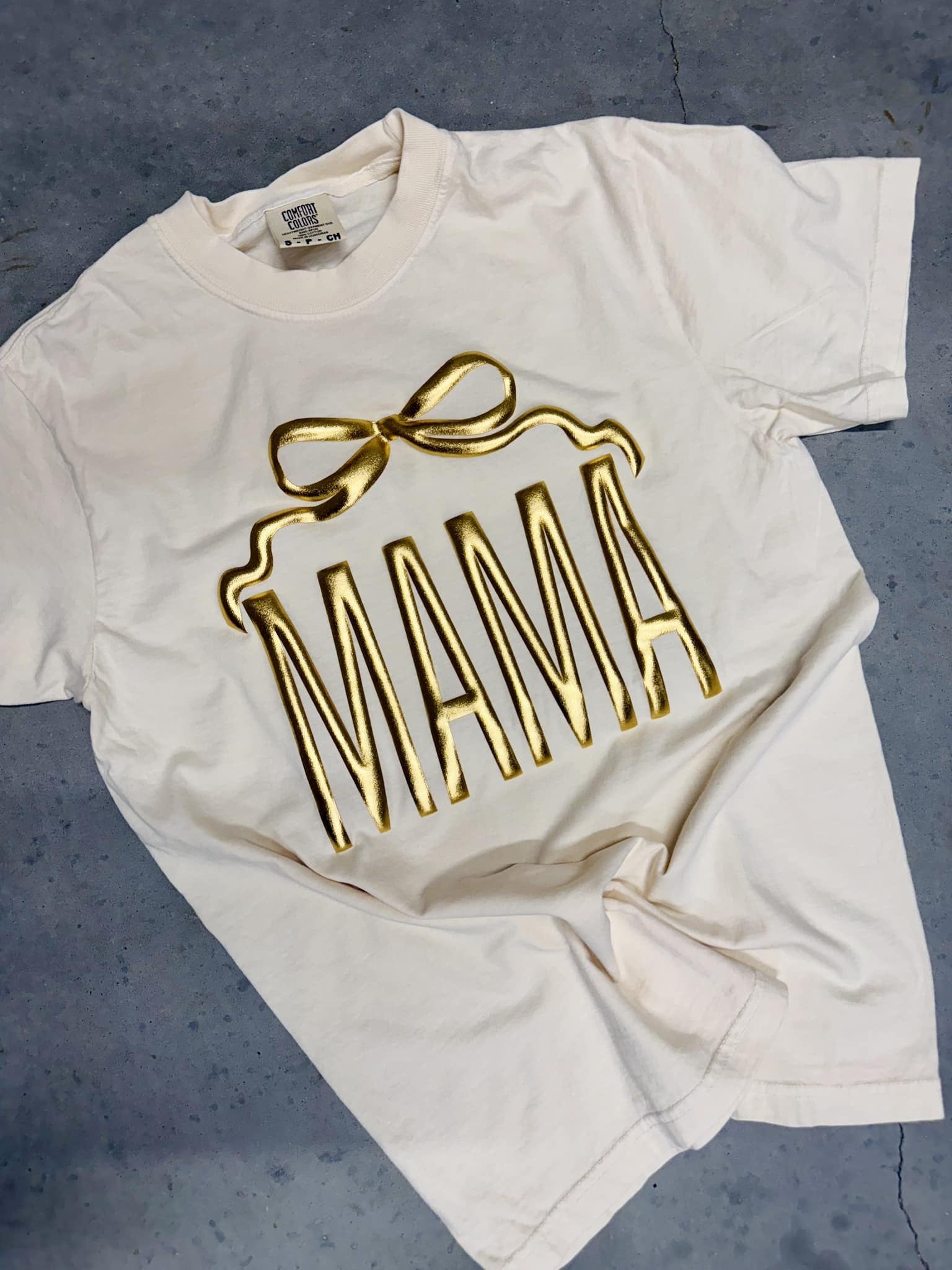 An ivory colored t-shirt with the word 'MAMA' printed in gold metallic vinyl in a bow shape.