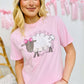 A person wearing a pink graphic tee with text and a sheep design, standing in front of a white background with decorative elements.
