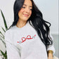 A woman smiling and sitting down, wearing a grey sweatshirt with a red and white embroidered bow design and the baseballs.