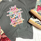 A gray t-shirt laid out on a patterned surface, featuring a printed design with the fruits of the spirit, accompanied by a pair of shoes.