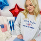 A woman sitting in front of a backdrop with American flag elements, wearing a gray sweatshirt with the text 'Land of the Free Home of the Cowboy' and stars.
