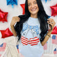 A person wearing a light blue graphic tee with a patriotic cherry design, shorts, and standing in front of a backdrop with red and blue stars.